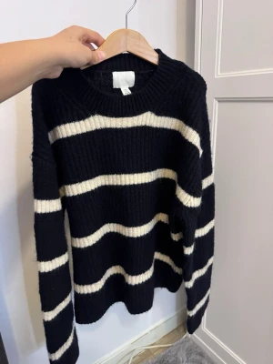 Svart stickad tröja med vita ränder - Mysig svart stickad tröja med breda vita horisontella ränder. Tröjan har rund halsringning, lång ärm och ribbstickade muddar. Perfekt för kalla dagar och enkel att matcha med jeans eller kjol.