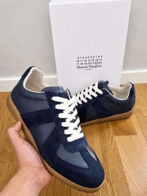 Maison Margiela GATs - Snygga sneakers från Maison Margiela i mörkblå mocka och läder med vita skosnören och beige gummisula. Klassisk low-top siluett som passar till många olika stilar. Märkeslogga på sulan och boxen ingår.