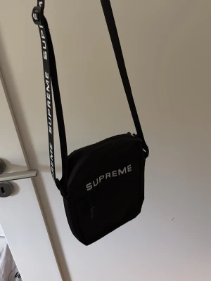 Svart Supreme axelväska - Säljer en svart axelväska från Supreme med justerbar axelrem och vit logga på både remmen och framsidan. Väskan har en fyrkantig form och dragkedja framtill. Perfekt för att bära det viktigaste och ger en streetwear vibe.