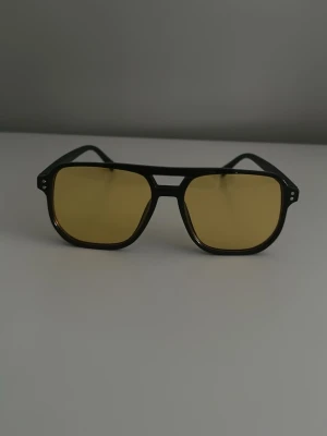 Svarta solglasögon med gula glas - Snygga solglasögon med svart, kantig båge och gula glas. Ramen har en klassisk pilotform och glasen ger en cool retrovibe. Perfekta för att sticka ut i sommar och ge din outfit en unik touch.