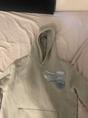 Beige hoodie med blå Mercier-tryck - Snygg beige hoodie från Mercier med flera blå och vita tryck både fram och bak. Klassisk huva och känguruficka framtill. Hoodien har en avslappnad passform och är gjord i mjukt material, perfekt för en chill och trendig look.