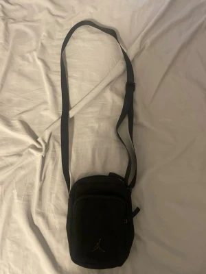 Svart axelväska med Jordan-logga - Snygg svart axelväska från Jordan med justerbar axelrem och två dragkedjefack. Väskan har en stilren design med den klassiska Jumpman-loggan framtill. Perfekt storlek för det viktigaste och enkel att bära crossbody.