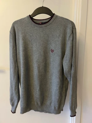 Grå stickad tröja med broderad logga - En stilren grå stickad tröja med rund hals och diskret broderad logga i vinrött på bröstet. Tröjan är i storlek M men stor i storleken så passa också L. Pris kan diskuteras vid snabb affär!