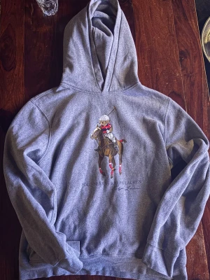 Grå hoodie med Polo Bear - Snygg grå hoodie från Polo Ralph Lauren med stort tryck av en polospelare på häst framtill. Klassisk huva och känguruficka. Mjuk bomullskänsla och avslappnad passform, perfekt för en chill och sportig look.
