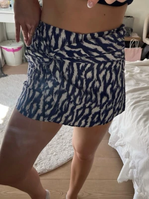 Blåvit mönstrad skort med knut - Supersnygg blåvit skort med trendigt abstrakt mönster och knutdetalj framtill. Skorten har en omlott-look och är tillverkad i ett mjukt, lätt tyg som känns skönt mot huden. Perfekt för dig som vill ha både kjol-look och shortskomfort.