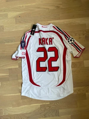 Kaka AC Milan 06/07 Bortatröja Matchtröja Retro Vintage - Säljer en vit AC Milan bortatröja från Adidas med röd och svart detaljering, Champions League-märke på ärmen och Kaka' 22 tryckt på ryggen. Tröjan har klubbmärke, sponsortryck och officiella patches. Perfekt för fotbollsfans!