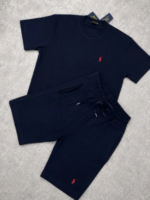 Mörkblått set Ralph Lauren - Snyggt mörkblått set från Polo Ralph Lauren med t-shirt och shorts. Båda plaggen har den klassiska röda logotypen broderad på bröstet respektive benet. Setet har en enkel och stilren design med rund halsringning och dragsko i midjan på shortsen. Perfekt för en avslappnad look.