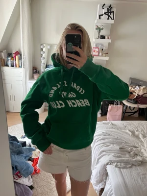 Grön Gant hoodie med tryck - Snygg grön hoodie från Gant med vit text 'Cedar Island Beach Club' på bröstet. Klassisk känguruficka framtill och stor huva. Perfekt för en avslappnad och sportig stil. Mjuk bomullskänsla och ribbade muddar vid ärmslut och nederkant.