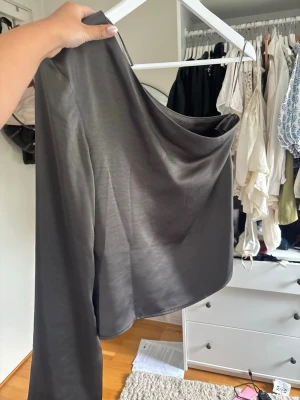Grå asymmetrisk topp med en ärm - Snygg grå topp i mjukt, glansigt material med en axel och lång ärm. Asymmetrisk design som ger en trendig look. Perfekt att styla med höga jeans eller kjol för en cool outfit.