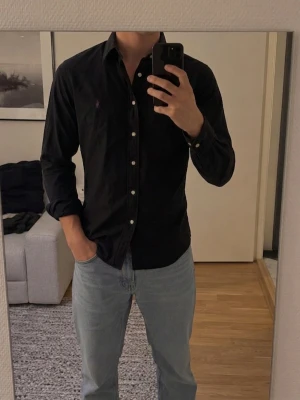Svart skjorta med knappar -  svart skjorta från Ralph Lauren med lila broderad logga på bröstet. Skjortan har lång ärm, knappar framtill och en smal passform. Tillverkad i bomull.Storlek M men är rätt så liten i passformen så skulle också kunna passa Någon som bär S.                    Skriv för mer information 😀