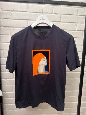Svart t-shirt med grafiskt tryck - Säljer en svart t-shirt med ett stort grafiskt tryck på bröstet som föreställer ett ansikte i profil med orange huva och svart bakgrund. T-shirten har rund hals och korta ärmar. Perfekt för dig som gillar unika prints och streetwear vibes.