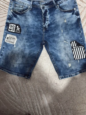 Blå jeansshorts med patchar - Säljer ett par blå jeansshorts med slitningar och coola patchar på framsidan. Shortsen har fem fickor, dragkedja och knapp, samt detaljer som tryck och lappar med text och mönster. Perfekta för en avslappnad streetstil.