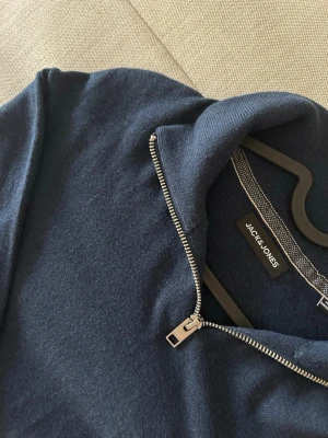 Mörkblå halvzip-tröja Jack & Jones - Mörkblå långärmad tröja från Jack & Jones med halvzip och hög krage. Tröjan har ribbade muddar vid ärmslut och nederkant, vilket ger en snygg och bekväm passform. Perfekt för dig som gillar stilrena och enkla plagg. Skriv för fler frågor!