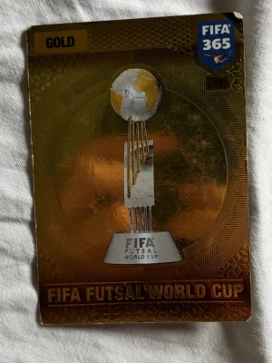 FIFA 365 Gold Futsal World Cup-kort - Samlarkort från FIFA 365-serien med motiv av FIFA Futsal World Cup-trofén. Kortet har en glänsande guldfärgad yta och detaljer i vitt och svart. Perfekt för dig som gillar fotboll och samlarkort.