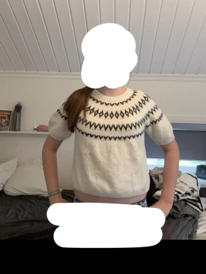 Vit stickad tröja med svart mönster - Kortärmad stickad tröja från Kappahl i vit färg med svart grafiskt mönster runt halsen. Tröjan har rund halsringning och ribbade kanter. Perfekt för lager på lager och ger en trendig look till din outfit.