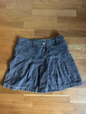 Grå manchesterkjol med fickor - Kort grå kjol i manchestertyg med bred linning, knappar och dragkedja fram. Kjolen har flera sydda veck och en liten ficka med broderad logga bak. Perfekt för dig som gillar en avslappnad men ändå trendig stil.