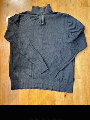 Mörkgrå stickad tröja Jack & Jones - Mörkgrå stickad tröja från Jack & Jones med halv dragkedja vid halsen. Tröjan har långa ärmar, ribbstickade muddar och nederkant. Perfekt för lager på lager och enkel att matcha med olika outfits.