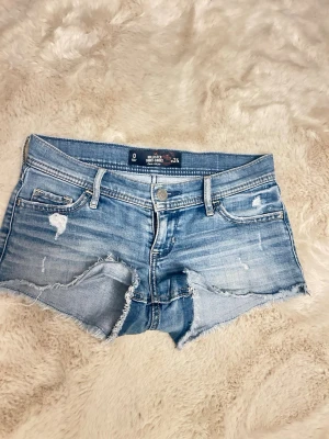 Low waist jeans shorts  - Säljer ett par ljusblå jeansshorts med slitna detaljer och råa kanter. Shortsen har fem fickor, bälteshällor och knappgylf. Perfekta för varma dagar och ger en avslappnad vibe. 