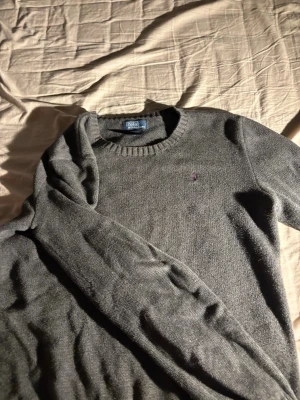 Grå stickad tröja från Polo Ralph Lauren - Mysig grå stickad tröja från Polo Ralph Lauren med rund halsringning och diskret broderad logga på bröstet. Tillverkad i mjuk bomull som passar perfekt för kyliga dagar. Klassisk och enkel design som funkar till det mesta.