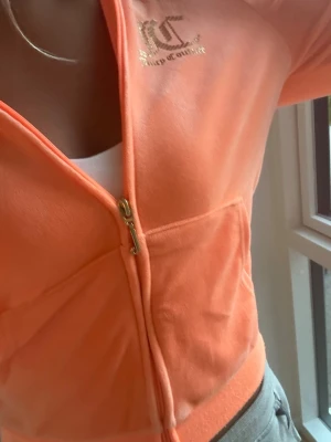 Persikofärgad Juicy Couture hoodie - Juicy Couture hoodie i mjuk velour, persikofärgad med guldfärgad logga på bröstet. Dragkedja framtill och fickor på sidorna. Perfekt för en chill och trendig look.(lite mindre i storleken)