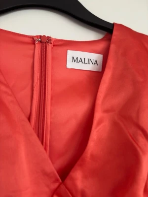 Röd kort klänning från Malina - Säljer en röd kort klänning från Malina i glansigt material. Klänningen har långa puffärmar med broderade detaljer och spets vid ärmslut och nederkant. Dragkedja bak och v-ringad hals. Perfekt för dig som vill sticka ut!
