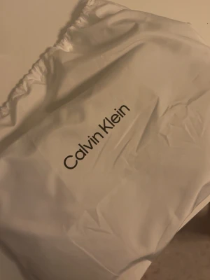 Svart quiltad kuvertväska Calvin Klein - Snygg svart kuvertväska från Calvin Klein med quiltad design och metallogo med CK på framsidan. Väskan har en stilren och modern look, perfekt till att lyfta vilken outfit som helst. Tillverkad i ett slätt material som ger en lyxig känsla.