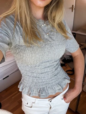 Grå smockad topp med volangkant - Superfin ljusgrå topp med smockad midja och volangkant nertill. T-shirtrmodell med korta ärmar och rund halsringning. Perfekt för en chill och trendig look.