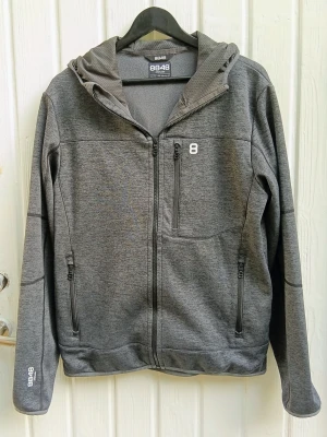 Hoodie från 8848 Altitude - Snygg grå hoodie från 8848 Altitude med dragkedja framtill, två sidofickor med dragkedja och en bröstficka. Tröjan har huva och diskreta loggor på bröstet och ärmen. Tillverkad i softshell, ett mjukt och stretchigt material som passar perfekt till träning.