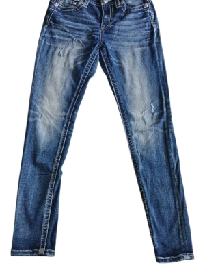 Miss Me Blå jeans med broderade fickor stl.28 - Säljer ett par blå jeans med snygga broderier på bakfickorna som föreställer ett tjurkranium och blommor. Jeansen har dekorativa nitar och kontrastsömmar, samt en patch med ett M på linningen. Perfekta för dig som gillar westernstil.