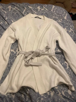 Vit omlottkofta med knytskärp - Snygg vit omlottkofta med knytskärp i midjan. Koftan har långa ärmar och en mjuk, stickad känsla som ger en avslappnad men ändå stilren look. Perfekt att slänga på sig över en topp för extra lager och stil.