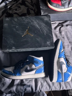  Air Jordan 1 Mid sneakers Signal blue - Snygga Air Jordan 1 Mid sneakers i blått och vitt med svarta detaljer. Skorna har klassisk siluett, vit tåbox och sidor, blå overlays och svart swoosh. Tillverkade i läder med svart yttersula och vita skosnören. Perfekta för dig som gillar streetwear och ikonisk design.