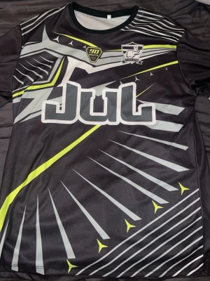 Svart sporttröja med grafiskt mönster - Säljer en svart sporttröja med gråa och neongula grafiska detaljer. Tröjan har rund halsringning och korta ärmar. Tryck med texten 'Jul' på bröstet och emblem på vänster sida. Tillverkad i ett lätt och snabbtorkande material, perfekt för träning.