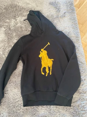 Svart hoodie med gul polospelare - Säljer en svart hoodie med stor gul polospelare broderad på bröstet. Hoodien har huva och ribbade muddar vid ärmar och nederkant. Perfekt för dig som gillar streetwear och vill sticka ut med en fet logga framtill.