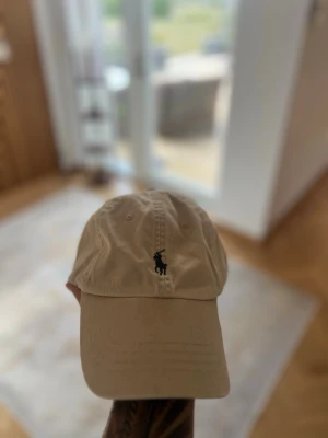 Beige keps från Polo Ralph Lauren - Snygg beige keps från Polo Ralph Lauren med klassisk broderad logga framtill och texten 'POLO' broderat baktill. Justerbart spänne bak för perfekt passform. Tillverkad i bomullstyg som andas och passar till de flesta outfits.