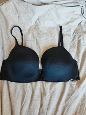 Svart bygel-bh från ELLA M - Snygg svart bh från ELLA M med bygel och vadderade kupor. Justerbara axelband och slät design utan spets eller mönster. Perfekt för dig som vill ha en enkel och stilren bh under alla typer av plagg.