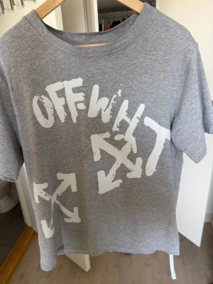 Grå Off-White t-shirt med tryck - Snygg grå t-shirt från Off-White med stort vitt tryck och grafiska pilar på framsidan samt logga och text på ryggen. Klassisk rund hals och korta ärmar. Perfekt för dig som gillar streetwear och vill sticka ut lite extra.