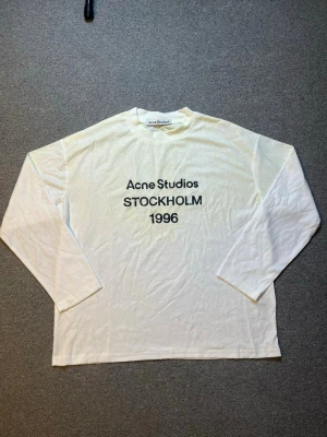 Vit långärmad topp Acne Studios - Vit långärmad topp från Acne Studios med tryck 'STOCKHOLM 1996' framtill. Klassisk rund halsringning och relaxed fit. Perfekt för dig som gillar minimalistisk och stilren design.