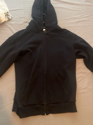 Svart zip-hoodie med fickor - En stilren svart hoodie med dragkedja framtill och stora fickor. Klassisk modell med huva och ribbade muddar vid ärmslut och nederkant. Perfekt för en avslappnad look och enkel att matcha med andra plagg.