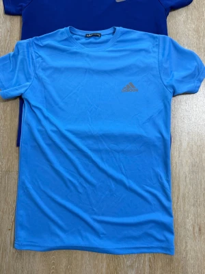 Blå t-shirt från Adidas - Snygg blå t-shirt från Adidas med klassisk logga på bröstet. T-shirten har rund halsringning och korta ärmar. Perfekt för en sportig och avslappnad stil.