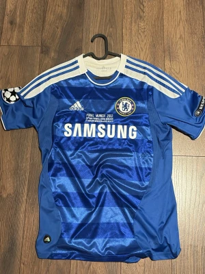 Chelsea FC Adidas matchtröja blå - Chelsea FC matchtröja från Adidas i blått med vita detaljer och tryck. Tröjan har Champions League-finalen 2012-tryck, klubbmärke, sponsorlogga och UEFA Respect-märke på ärmen. Tillverkad i lätt och ventilerande material, perfekt för fotboll eller samling.