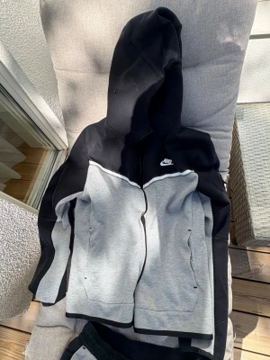 Nike svart och grått mysdress-set - Snyggt set från Nike med zip-hoodie och matchande byxor i svart och grått. Hoodien har dragkedja, fickor och vit Nike-logga på bröstet. Byxorna har dragsko i midjan och logga på låret. Perfekt för en avslappnad stil.