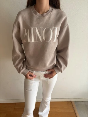 Beige sweatshirt med MINOT-tryck - Säljer en beige sweatshirt med stort vitt MINOT-tryck på bröstet. Tröjan har rund halsringning, avslappnad passform och ribbade muddar vid ärmslut och nederkant. Tillverkad i mjukt bomullsliknande material, perfekt för en chill och stilren look.