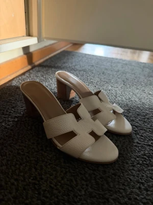 Vita slip-in sandaler med klack - Säljer ett par stilrena vita slip-in sandaler med breda remmar i skinnimitation och öppen tå. Sandalerna har en chunky träfärgad klack och ett trendigt utskuret mönster på ovandelen. Perfekta för dig som vill ha en enkel men snygg look till sommaren.