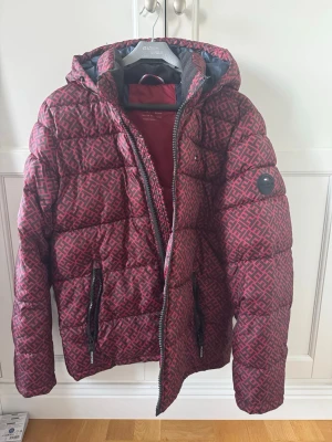 Röd dunjacka från Tommy Hilfiger - Snygg röd dunjacka från Tommy Hilfiger med svart mönster, huva och dragkedjor. Jackan har två framfickor med dragkedja och en innerficka. Perfekt för kalla vinterdagar och har en modern, sportig look.