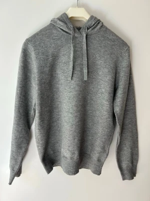 Grå stickad hoodie med huva - En stilren grå stickad hoodie med huva och dragsnören. Tröjan har ribbade muddar vid ärmslut och nederkant, samt en mjuk och bekväm känsla. Perfekt för dig som gillar en enkel men snygg look. Hör av dig om du har några frågor!