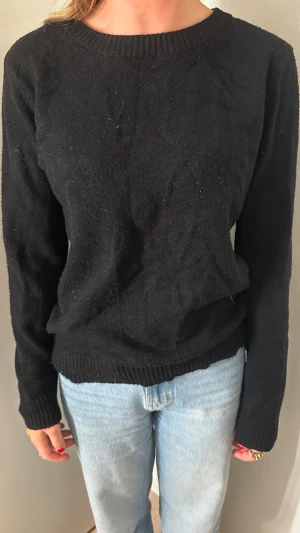 Svart stickad tröja med rund hals - En enkel och stilren svart stickad tröja med rund halsringning och långa ärmar. Tröjan har ribbade muddar vid ärmslut och nederkant. Perfekt att matcha med jeans för en avslappnad look.