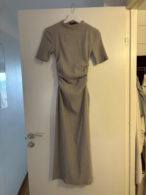 Beige klänning från Zara - Säljer en beige klänning med korta ärmar. Klänningen har en figurnära passform med snygg rynkning i midjan som ger en cool siluett. Materialet är mjukt och stretchigt, perfekt för en trendig look.