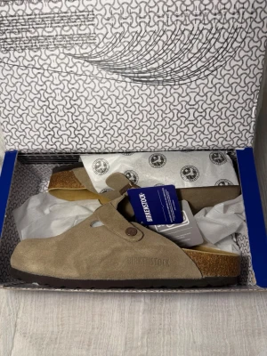 Taupe mocka från Birkenstock - Säljer ett par snygga beige tofflor från Birkenstock i mocka med justerbart spänne och korksula. Perfekta för dig som vill ha både stil och komfort. Yttersulan är i gummi och innersulan är formad för extra stöd. ‼️Skickar inom 24H‼️ **PRIS KAN DISKUTERAS**