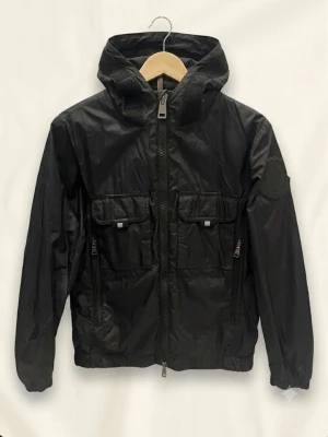 Moncler Windbreaker Vingeanne Giubbotto 152cm - Säljer en stilren och äkta Moncler-jacka i klassiskt svart modell Vingeanne Giubbotto. Perfekt som tunn vår-, höst- eller sommarjacka! • Märke: Moncler (100% äkta, se koden på etiketten: F19541A70320) • Storlek: 12 Anni / 152 cm (Passar även en mindre XS) • Detaljer: Gummerad Moncler-logga på ärmen, loggodetalj längs huvan, bröstfickor och praktiska fickor med dragkedja. • Skick: Mycket fint och välvårdat skick. 📦 Snabbskickat och noggrant paketerat! Hör av dig om du har frågor!
