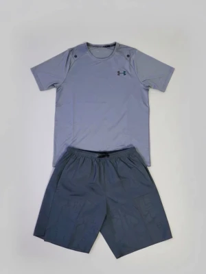 Grått träningsset från Under Armour - Snyggt grått träningsset från Under Armour med en kortärmad t-shirt och matchande shorts. Tillverkat i ett lätt och snabbtorkande material som passar perfekt för gymmet eller löprundan. Diskret logga på bröstet och elastisk midja på shortsen.